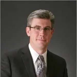 Professional head-and-shoulders portrait of a man in a dark suit, tie, and glasses against a gray backdrop.