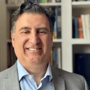 Smiling man in a gray suit and light blue shirt, portrait in front of a bookshelf.