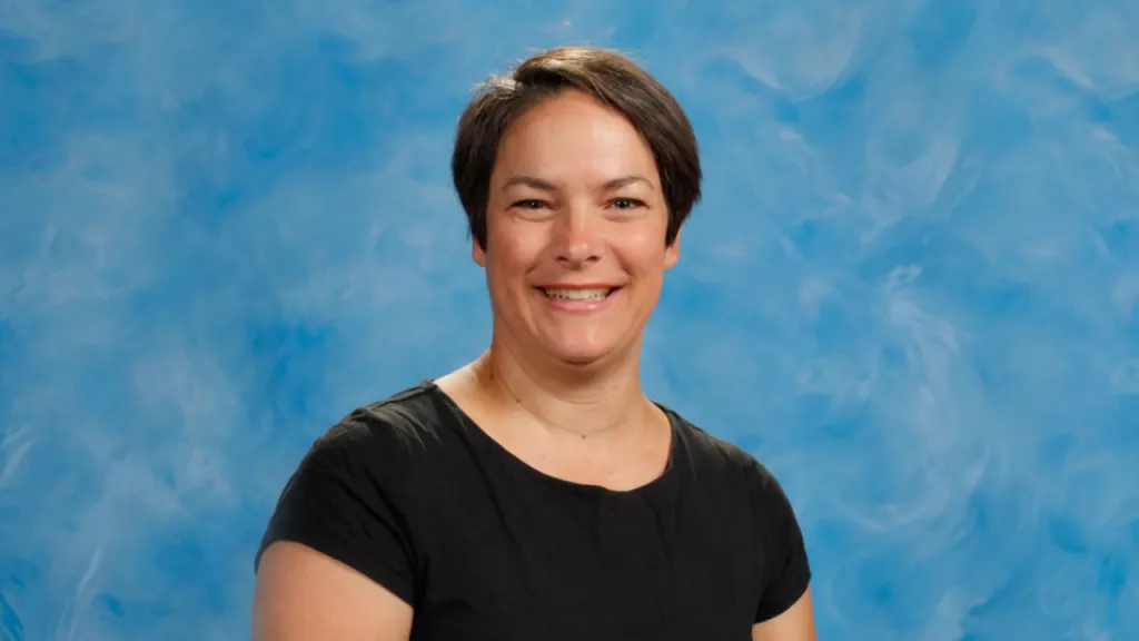 Smiling woman with short dark hair wearing a black shirt, posed in a blue studio backdrop portrait.