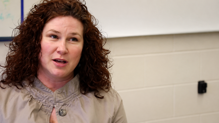 Woman with curly brown hair speaks, wearing a beige jacket in a classroom setting.