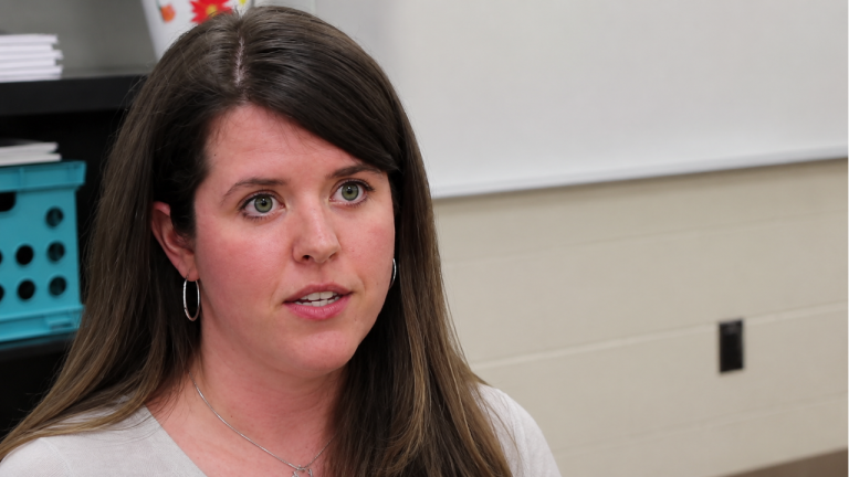 Close-up of a woman with brown hair and hoop earrings speaking in a classroom setting, neutral expression, whiteboard behind her.