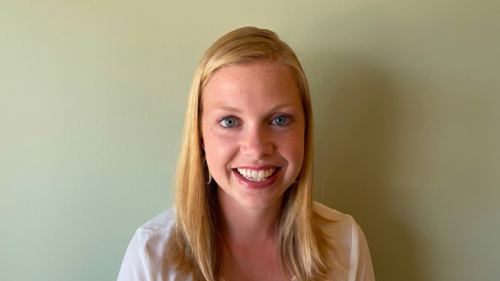 Smiling young woman with shoulder-length blonde hair standing against a pale green wall.