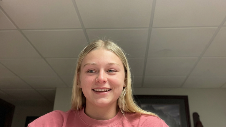Young woman with blonde hair wearing a pink sweatshirt, smiling in an indoor room with a grid ceiling behind her.