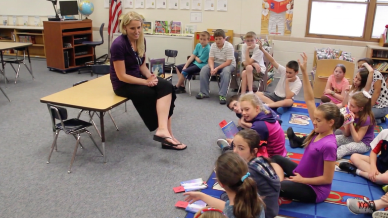 Teacher sits on a desk at the front of a classroom as students on the carpet listen, some raising hands.