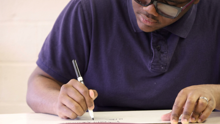 Close-up of a person in a purple polo shirt wearing sunglasses, writing with a marker on a white sheet at a table.