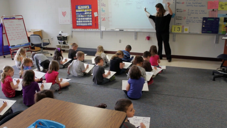 Teacher standing at whiteboard teaching a group of children seated on the carpet, each child writing on small white boards or paper pads.