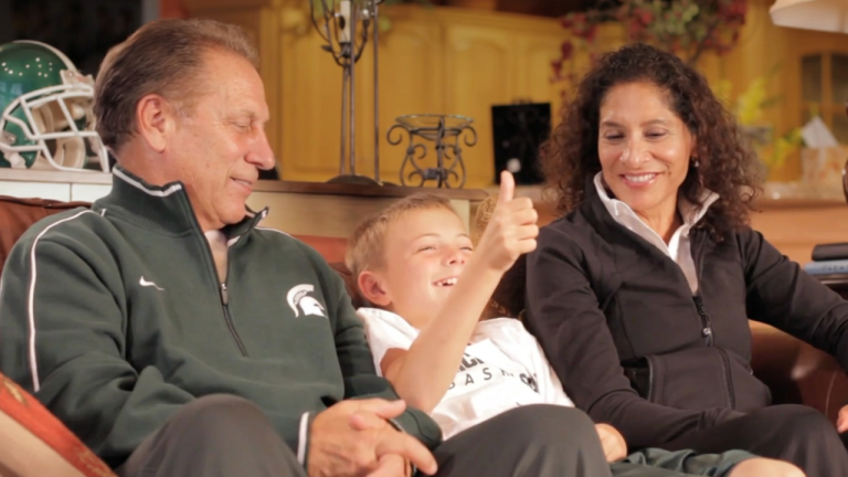 Three people sitting on a living-room couch: an older man in a green jacket, a smiling boy giving a thumbs up, and a woman in a black jacket smiling nearby.
