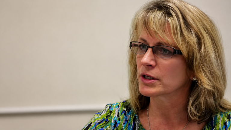 Woman with shoulder-length blonde hair and glasses speaking, wearing a green patterned blouse in an indoor setting of a meeting or interview.