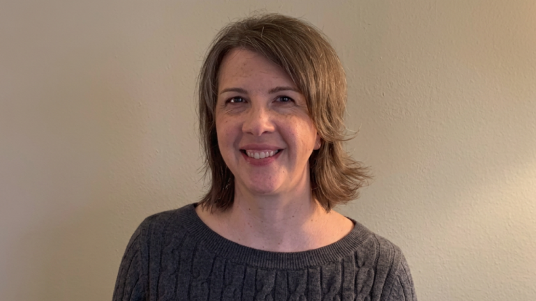 Portrait of a smiling woman with short brown hair wearing a gray knitted sweater against a beige wall.