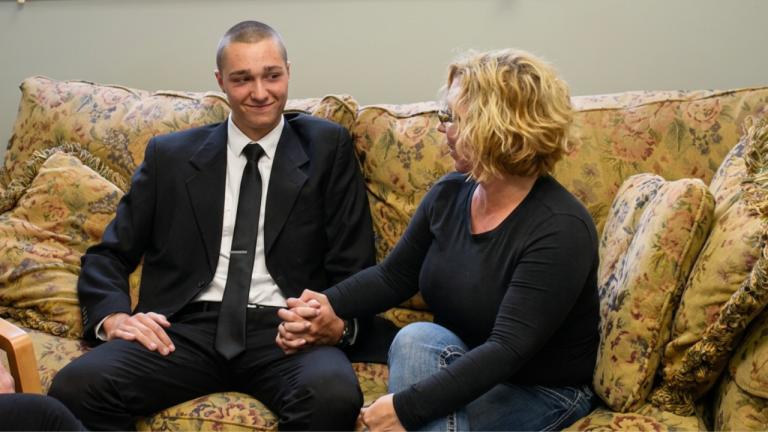 Young man in a suit and tie sits on a floral couch, holding hands with a woman in a black long-sleeve shirt and jeans