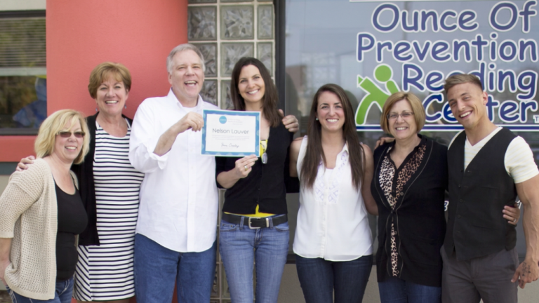 Group of seven adults posing outside a reading center, with one man in a white shirt holding a certificate and smiling.