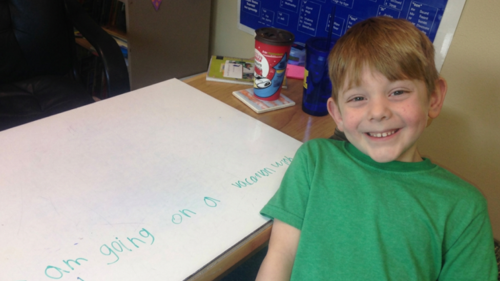 Smiling boy in a green shirt sits at a desk with a whiteboard, which has teal writing on it in cursive. Behind him are classroom supplies and a blue water bottle.