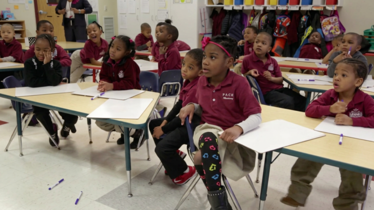 A classroom of young students in maroon uniforms sit at desks with papers and markers, listening to a teacher.