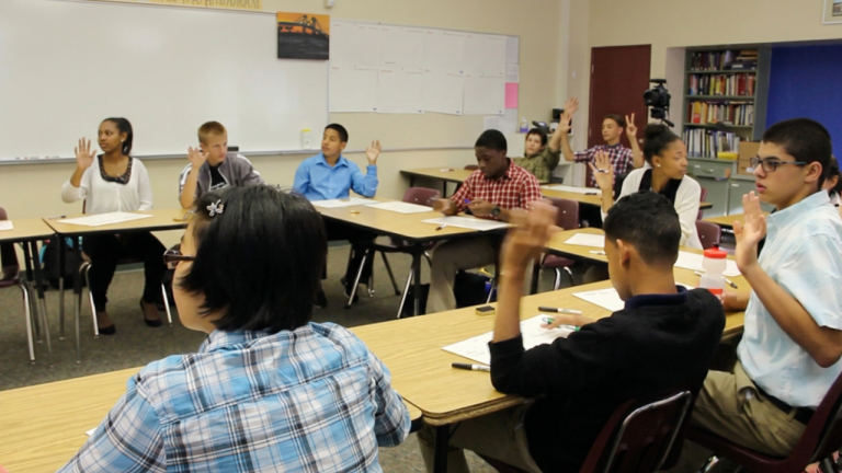 Diverse students seated around U-shaped desks in a classroom, many raising their hands during a discussion, with a whiteboard and bookshelves in the background.