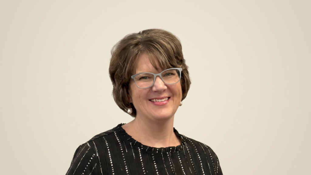 Smiling woman with short brown hair and gray glasses, wearing a black striped blouse and pearl earrings against a beige background.