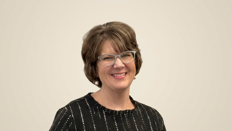 Smiling woman with short brown hair and gray glasses, wearing a black striped blouse and pearl earrings against a beige background.
