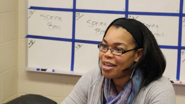 Woman with glasses and a scarf speaks, seated in front of a calendar board that shows 'Spring Break' entries behind her.