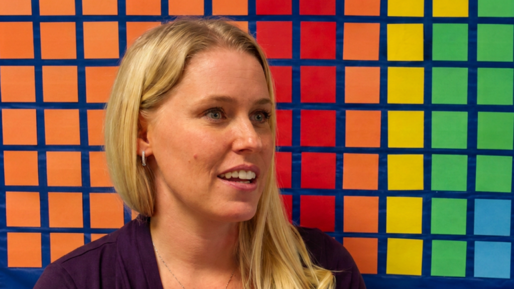 Close-up of a smiling blonde woman in a purple top, with a bright multicolored square grid background.