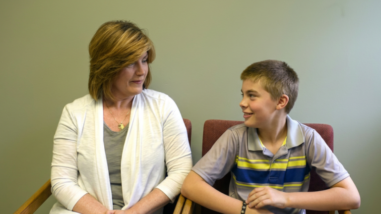 Woman and a boy seated next to each other on chairs, smiling as they talk in a room with a green wall.