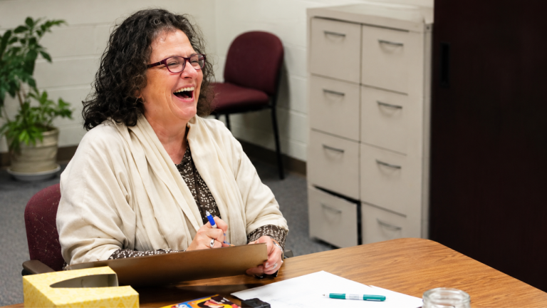 Smiling woman with curly hair and glasses laughs while writing on a clipboard at a wooden desk in an office.