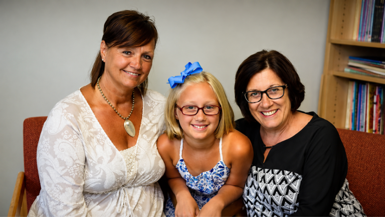Three smiling people—two adults and a young girl—pose on a red couch with bookshelves in the background.