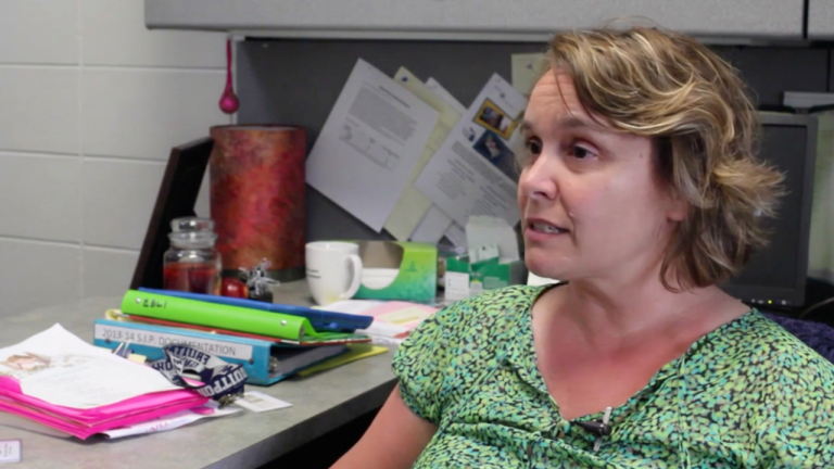 Woman in a green patterned blouse sits at a cluttered desk with scattered papers, folders, and a mug as a computer monitor looms in the background.